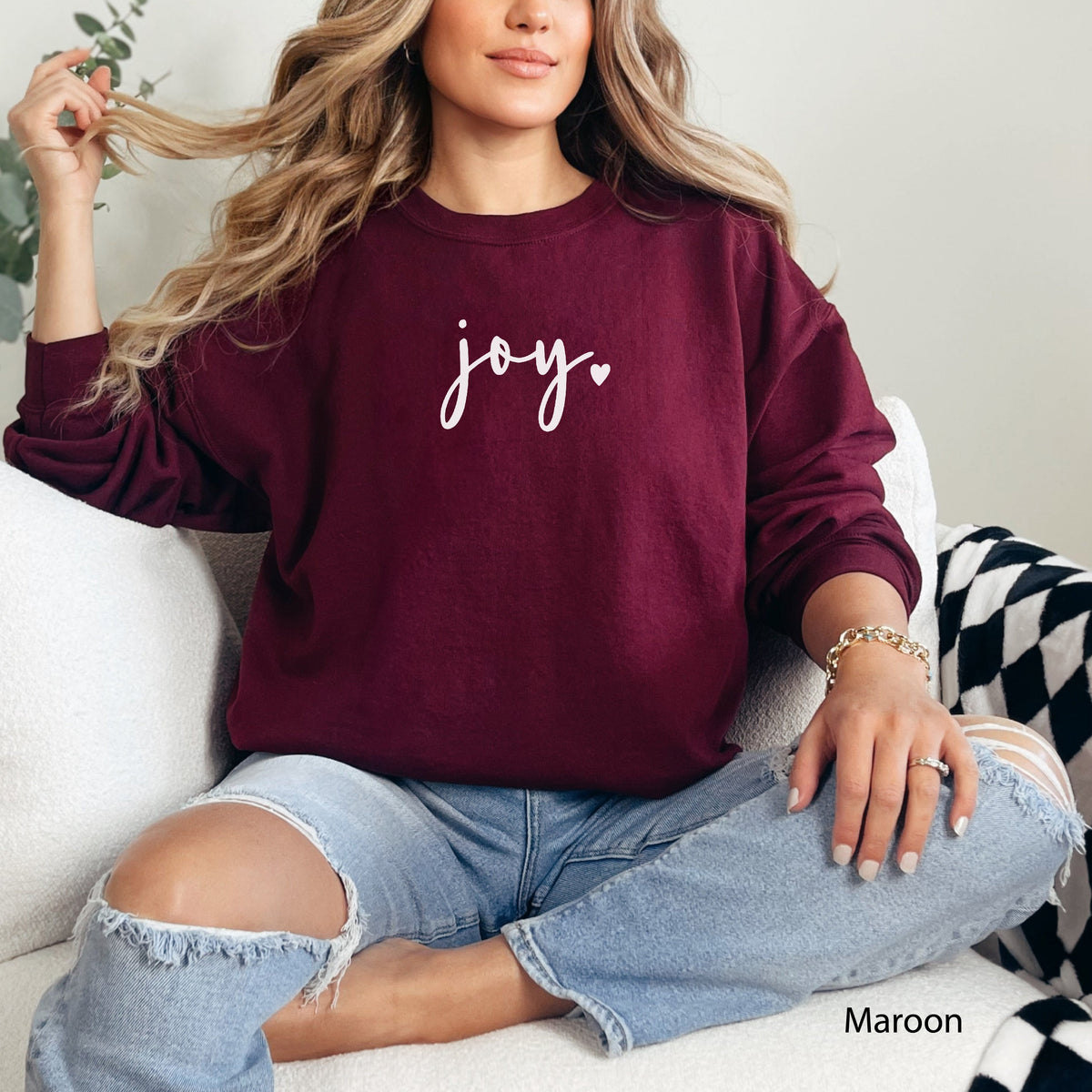 Maroon: Woman wearing a maroon Joy heart cursive graphic sweatshirt sitting on a white couch