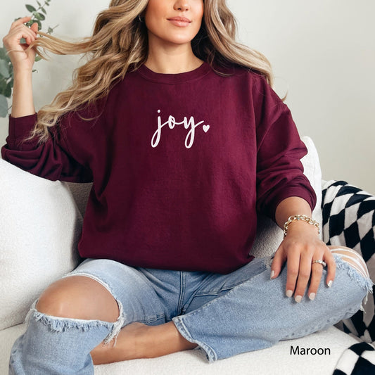 Maroon: Woman wearing a maroon Joy heart cursive graphic sweatshirt sitting on a white couch